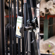 A smartphone is secured with the Mega Grip™ Forklift Front Guard Mount clamp holder. The phone displays a spreadsheet, set against industrial pipes, bolts, and rolled materials for an authentic warehouse backdrop.