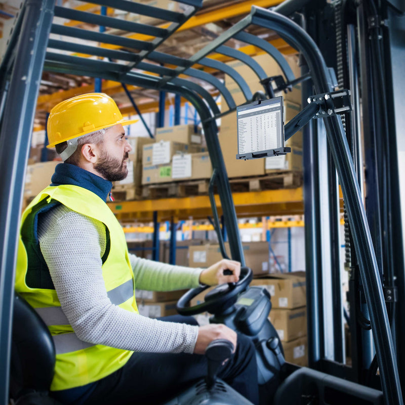 A man in a yellow safety vest and hard hat operates a forklift inside a warehouse, using the LockVise™ Metal Locking Forklift Front Guard Tablet Mount with 3.75 Shaft to view information on the screen. Boxes are stacked on shelves in the background.