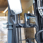 The LockVise™ Metal Locking Forklift Front Guard Tablet Mount with 5 Metal Robust™ Shaft secures a tablet near the steering wheel, showing a digital interface. Bolts, a rearview mirror, and stacked warehouse boxes are visible in the background.