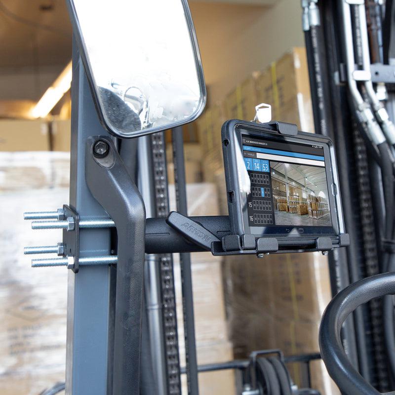 The LockVise™ Metal Locking Forklift Front Guard Tablet Mount with 5 Metal Robust™ Shaft secures a tablet near the steering wheel, showing a digital interface. Bolts, a rearview mirror, and stacked warehouse boxes are visible in the background.