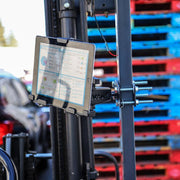 A tablet secured by the LockVise™ Metal Locking Forklift Front Guard Tablet Mount with 5 Metal Robust™ Shaft displays industrial data charts. Outdoors, stacks of red and blue pallets and blurred vehicles appear in sunlight.