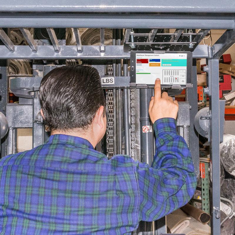 A person in a blue plaid shirt operates a forklift and uses the touchscreen display secured by the LockVise™ Metal Locking Forklift Overhead Guard Tablet Mount with 3.75 Shaft in a busy warehouse.