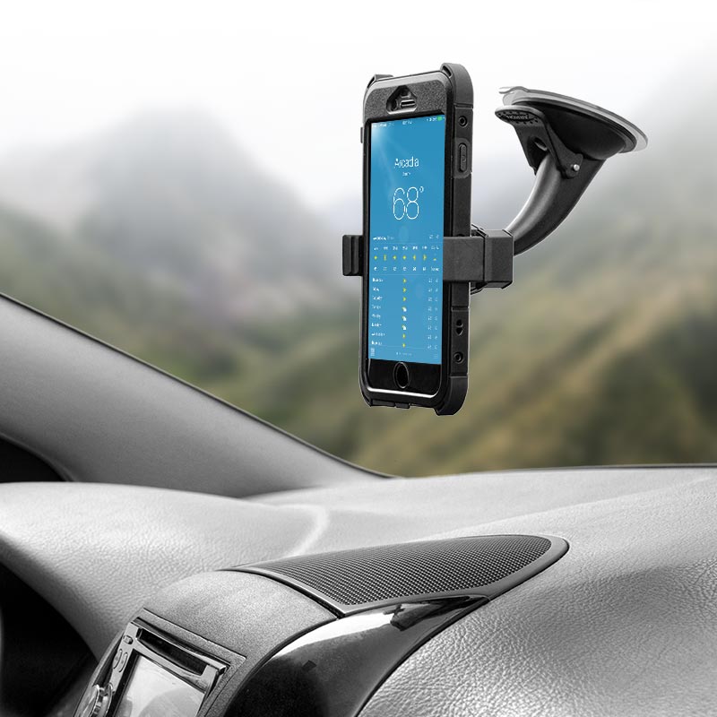 A smartphone in a black case is secured in a Mobile Grip 2 Holder with Suction Mount on the car dashboard, displaying a weather app, with the car interior and blurred mountains visible in the background.