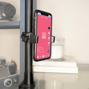 A smartphone is mounted vertically in the Mobile Grip 5 Phone Holder with Clamp Mount, attached to a pole. The screen shows a social media app with a pink background; cameras, a white candle, and stacked books are visible behind it.