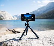 A RoadVise® XL 11 inch Tripod Mount Phone and Midsize Tablet Holder is set on a rock, capturing video or photos of a scenic lake with mountains and trees under a blue sky—ideal for creating content in stunning outdoor locations.