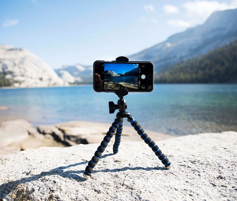 A RoadVise® XL 11 inch Tripod Mount Phone and Midsize Tablet Holder is set on a rock, capturing video or photos of a scenic lake with mountains and trees under a blue sky—ideal for creating content in stunning outdoor locations.