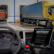 The LockVise™ Metal Locking Tablet Mount with Security Bundle secures an Android tablet displaying graphs to a trucks dashboard, with the steering wheel in view and several large trucks visible outside by a warehouse loading dock.