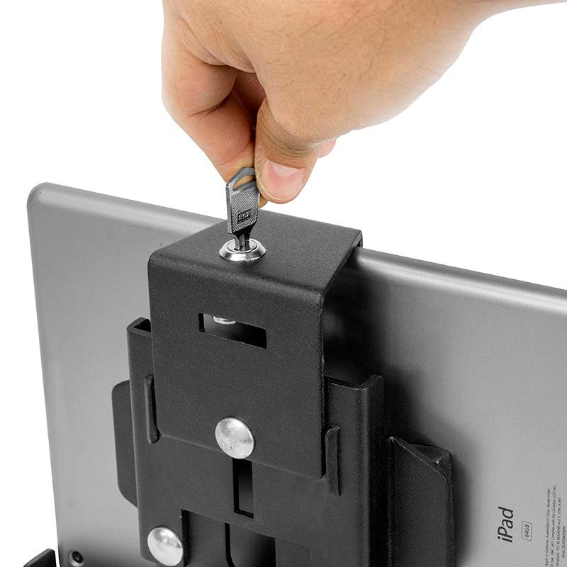 A hand inserts a key into the LockVise™ Metal Locking Tablet Mount, which is attached to the back of a gray iPad. The mount features a drill base and includes a magnetic micro USB charge cable for commercial use.