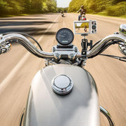 POV from a motorcycle, featuring the Premium Aluminum Motorcycle Handlebar Camera Mount recording two riders ahead. The shot includes handlebars, speedometer, and trees set against a sunny blue sky.