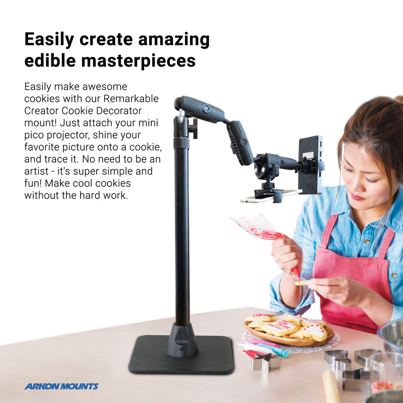 A woman uses the Remarkable Creator™ Cookie Decorators Mount Stand for Phones and Pico Projectors to project images onto cookies for tracing, surrounded by decorated cookies and tools, wearing a pink shirt and blue apron.