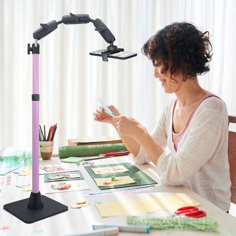 A woman crafts at a table, recording her work with the Remarkable Creator™ Pro Mount with Pink Extension Pole. The setup, bathed in sun through sheer curtains and surrounded by supplies, is ideal for any content creator.