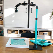 The Remarkable Creator™ Pro Mount for Phone or Camera with a teal extension pole is set up on a wooden desk, beside a framed teal print and folded cloth. Shelves holding baskets and decor are visible in the background.