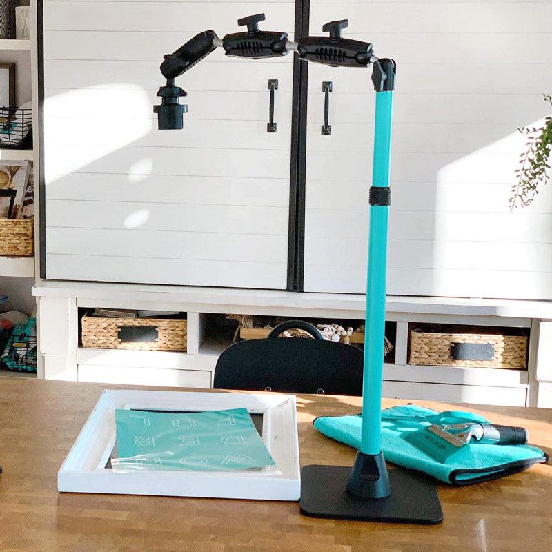 The Remarkable Creator™ Pro Mount for Phone or Camera with a teal extension pole is set up on a wooden desk, beside a framed teal print and folded cloth. Shelves holding baskets and decor are visible in the background.
