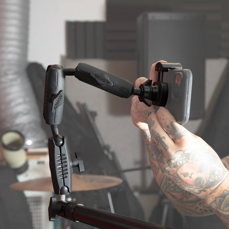 A tattooed person adjusts a smartphone held by the Remarkable Creator™ Pro Stand with Clamp Base in a music studio, with blurred musical instruments in the background.