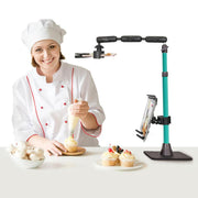 A smiling chef decorates cupcakes while recording with a smartphone mounted on the Remarkable Creator™ Pro+Plus with Teal Extension Pole, which captures perfect overhead shots of the content creation process.