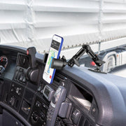 A smartphone on a truck dashboard displays a navigation map, secured by the RoadVise Heavy-Duty Multi-Angle Phone Mount (4-Hole AMPS Compatible), with the steering wheel and window showing a large truck parked outside.
