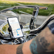 A smartphone with GPS is secured in a RoadVise® Holder with Chrome Aluminum Mount on motorcycle handlebars, as a tattooed rider grips the bike against a backdrop of green grass and pavement.