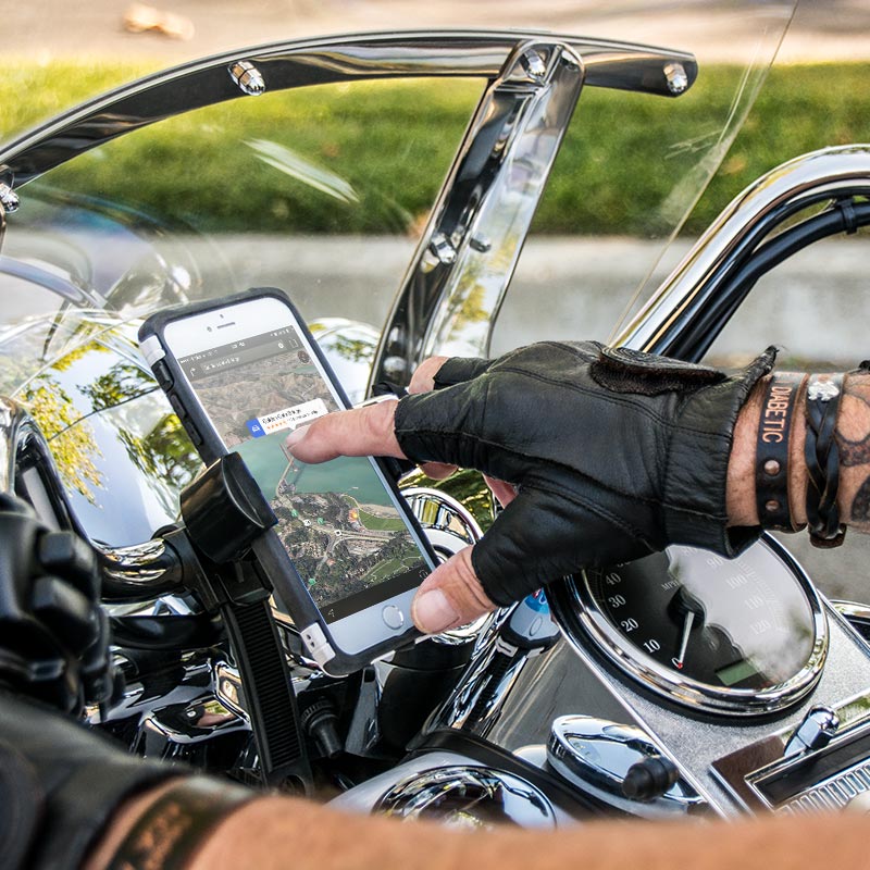 A person wearing black leather gloves uses a GPS app on a smartphone secured in a RoadVise® Motorcycle Phone Mount with Strap attached to the handlebars of a motorcycle, with the dashboard and chrome details visible.
