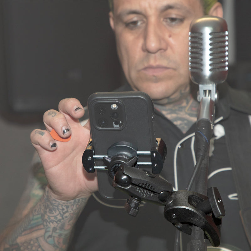A person with tattooed arms and painted nails adjusts a smartphone on the RoadVise® Robust Clamp Phone Mount with Security Knob near a vintage-style microphone, appearing focused as the phone faces them.
