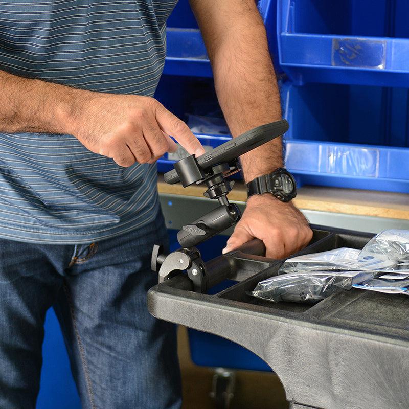 A person in a striped shirt and watch adjusts the RoadVise® Robust Clamp Phone Mount with Security Knob on a black arm attached to a utility cart, near blue storage bins and plastic-wrapped items.