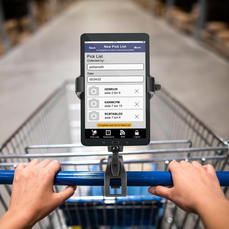A person pushes a shopping cart equipped with the RoadVise® Ultra Phone and Tablet Holder with Clamp Mount and Security Knob, displaying a pick list for collecting items in a warehouse or store aisle.