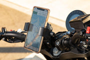 A smartphone with a map is held by the RoadVise® Ultra Holder with Aluminum Handlebar Mount on motorcycle handlebars for safe navigation. The photo is taken outdoors in daylight.
