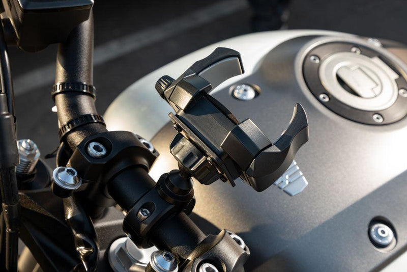 Close-up of a motorcycle handlebar fitted with the RoadVise® Ultra Holder with Aluminum Handlebar Mount. Sunlight gleams off the bike’s metallic fuel tank, visible fuel cap, and Suzuki logo in the background.