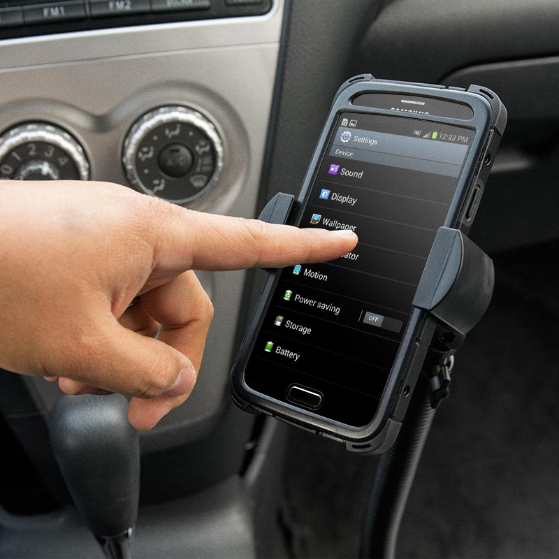 A persons hand touches a smartphone screen on a car dashboard using a RoadVise® XL Car Seat Rail or Floor Mount. The phone shows its settings menu, and car controls appear in the background.