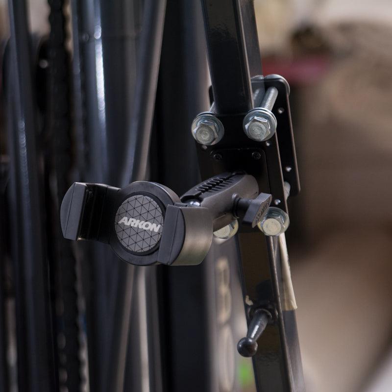 Close-up of the RoadVise® XL Forklift Front Guard Phone and Midsize Tablet Mount attached to a black metal frame with bolts, featuring its adjustable arm and grip. The blurred background highlights this sturdy warehouse device mount.