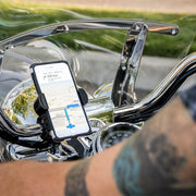 A RoadVise® XL Motorcycle Phone Mount - Black Aluminum secures a smartphone on the handlebars, GPS visible on screen. A tattooed arm appears in front, with a shiny windshield and chrome details gleaming behind.