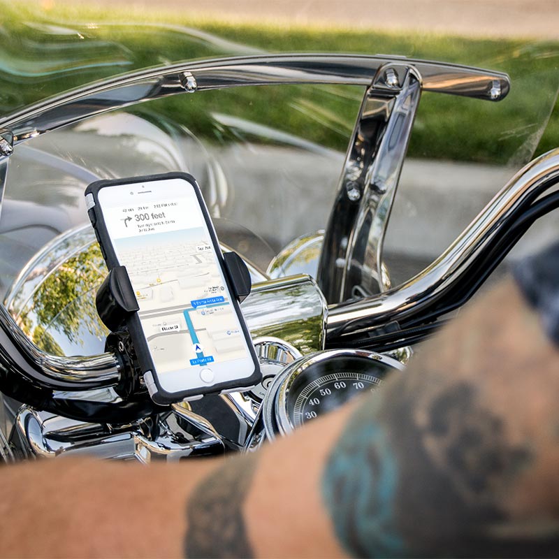 A RoadVise® XL Motorcycle Phone Mount - Black Aluminum secures a smartphone on the handlebars, GPS visible on screen. A tattooed arm appears in front, with a shiny windshield and chrome details gleaming behind.