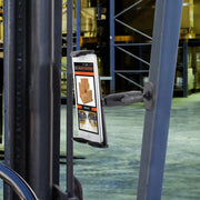 The Robust Magnetic Tablet Mount securely attaches a tablet to a forklift, showing inventory info and box photos, with warehouse shelves full of goods visible in the background.