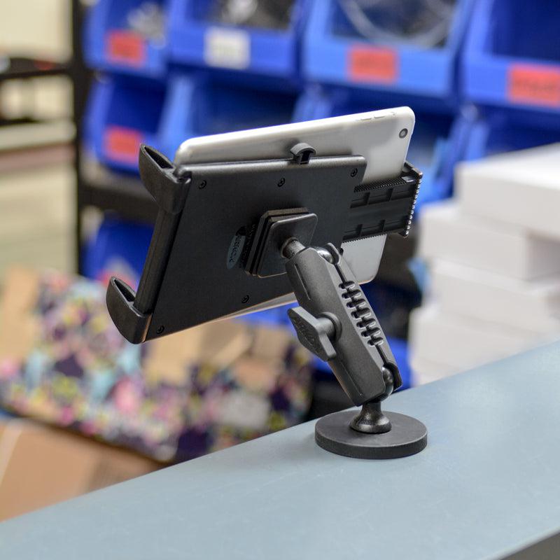 The Robust Magnetic Tablet Mount, similar to a forklift tablet holder, is attached to a desk edge holding a tablet securely. In the background, blue storage bins and stacked boxes can be seen in a busy workspace.