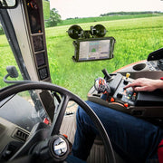 A person operates a modern tractor using control panels and a touchscreen, green fields visible outside, while a Slim-Grip® Double Suction Windshield Tablet Mount secures an extra screen to the dashboard.