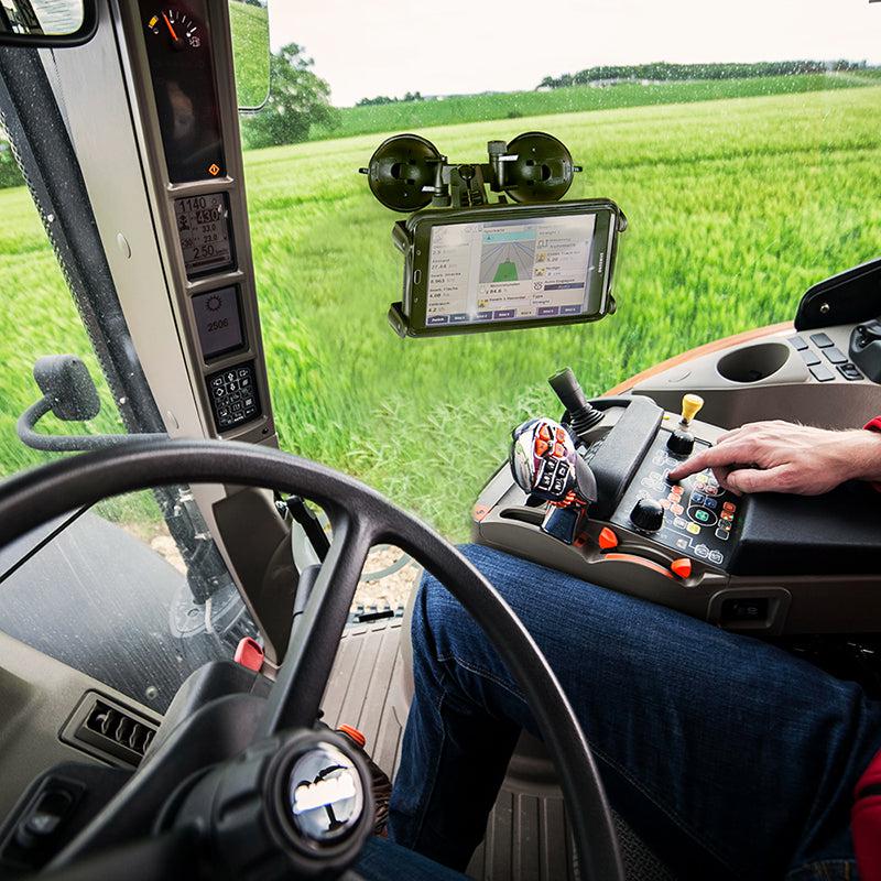 A person operates a modern tractor using control panels and a touchscreen, green fields visible outside, while a Slim-Grip® Double Suction Windshield Tablet Mount secures an extra screen to the dashboard.