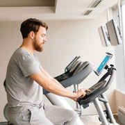A man in grey workout clothes uses a stationary bike in a gym, viewing a screen secured by the Slim-Grip® Tablet Clamp Mount attached to the handlebars. Other gym equipment and large windows can be seen in the background.