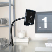 The Slim-Grip® Tablet Holder with Clamp Mount and 22 Gooseneck is attached to a desk near a shelf of cameras, next to a monitor showing 1 PM and a candle in a glass holder.