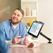 A smiling healthcare professional in scrubs sits at a desk, holding a pen and talking to someone. A heart diagram is displayed on a tablet held by the Slim-Grip® Tablet Holder with Clamp Base and Extension Arm in front of him.