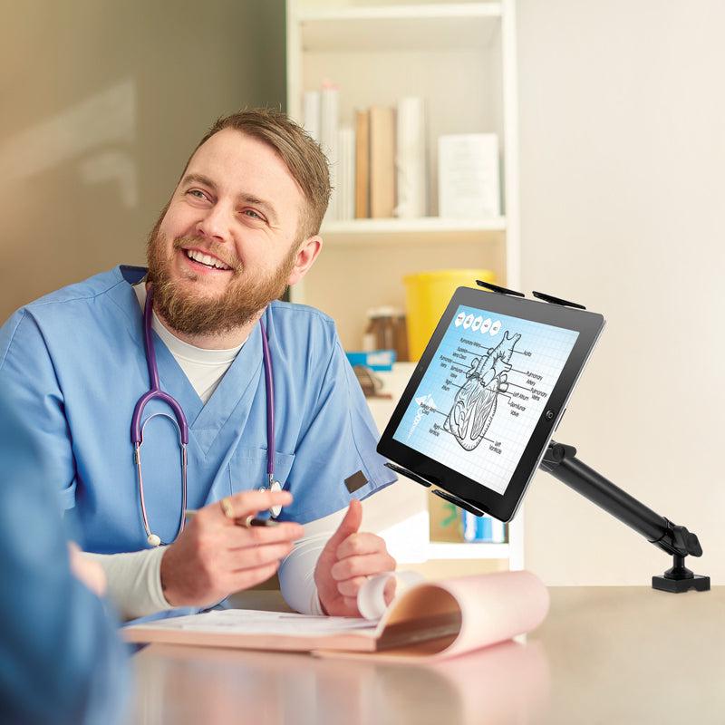 A smiling healthcare professional in scrubs sits at a desk, holding a pen and talking to someone. A heart diagram is displayed on a tablet held by the Slim-Grip® Tablet Holder with Clamp Base and Extension Arm in front of him.