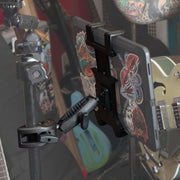 A tablet covered in colorful stickers is held by the Slim-Grip® Tablet Holder with Clamp Mount and Security Knob Shaft, attached to a stand, with a green electric guitar and other instruments blurred in the background.