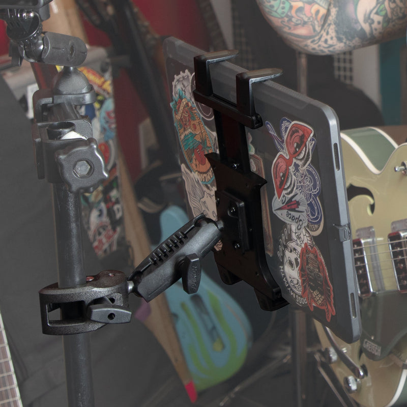 A tablet covered in colorful stickers is held by the Slim-Grip® Tablet Holder with Clamp Mount and Security Knob Shaft, attached to a stand, with a green electric guitar and other instruments blurred in the background.