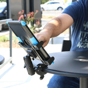 A person in a blue shirt adjusts a tablet secured in a Slim-Grip® Tablet Holder with Clamp Mount and Security Knob Shaft, which is attached to the edge of a round outdoor table. Plants, cars, and buildings are blurred in the background.