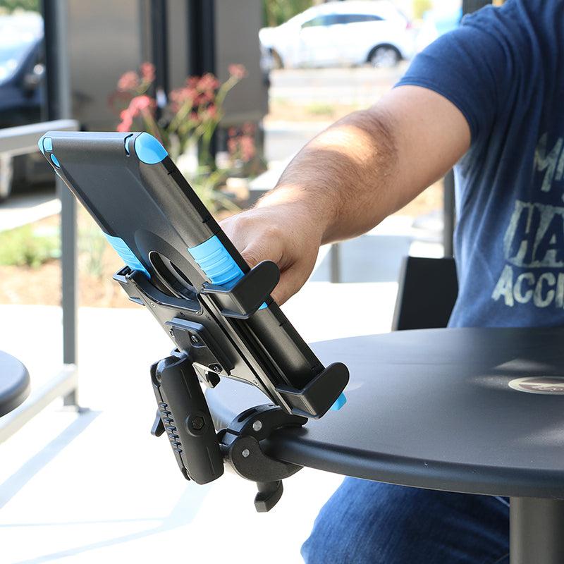 A person in a blue shirt adjusts a tablet secured in a Slim-Grip® Tablet Holder with Clamp Mount and Security Knob Shaft, which is attached to the edge of a round outdoor table. Plants, cars, and buildings are blurred in the background.
