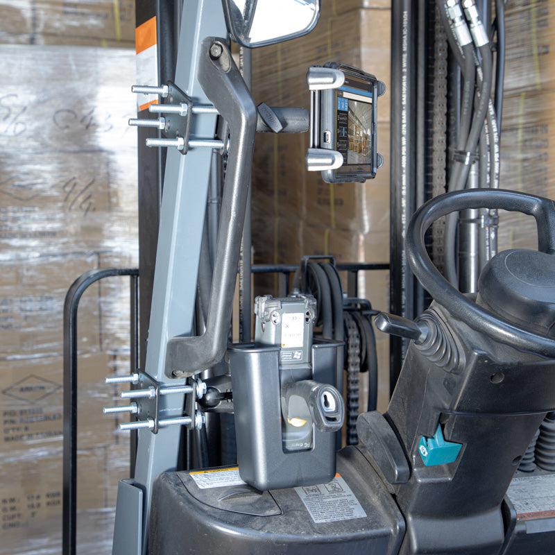 A close-up shows a forklift control area with a steering wheel, levers, and the Slim-Grip® Tablet Holder with Forklift Front Guard Mount and 3.75 Shaft, plus a barcode scanner; stacked boxes on pallets fill the background.