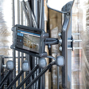 The Slim-Grip® Tablet Holder with Forklift Front Guard Mount and 3.75 Shaft is attached to a metal frame near control levers, showing warehouse inventory data, with pallets wrapped in plastic seen in the background.
