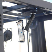 A Slim-Grip® Tablet Holder with Forklift Overhead Guard Mount and 6 Metal Shaft secures a smartphone upside down on a black metal bracket attached to a steel frame, with blurred industrial equipment in the background.