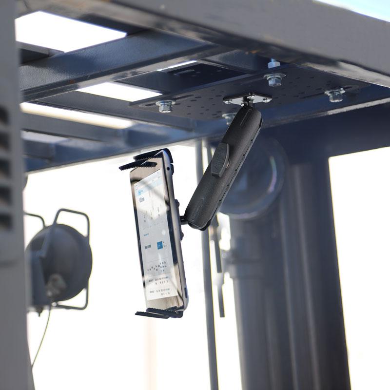 A Slim-Grip® Tablet Holder with Forklift Overhead Guard Mount and 6 Metal Shaft secures a smartphone upside down on a black metal bracket attached to a steel frame, with blurred industrial equipment in the background.