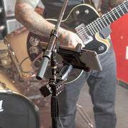 A tattooed musician plays an electric guitar by a drum set. In front of them, a Slim-Grip® Tablet Holder with Heavy-Duty Clamp Mount secures a tablet on a stand, likely displaying music or lyrics in the rehearsal space.
