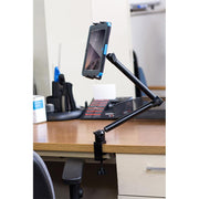 A tablet is held upright in the Slim-Grip® Tablet Holder with Heavy-Duty Desk or Wheelchair Clamp Mount and 22 Arm, which is securely attached to the edge of a wooden desk in an office with various supplies in the background.