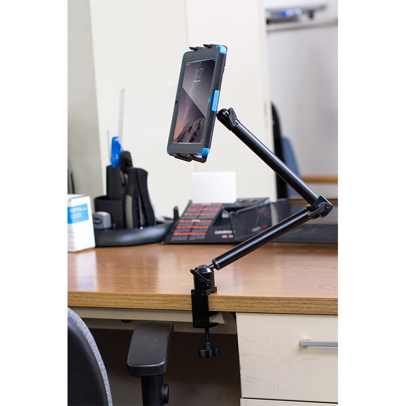 A tablet is held upright in the Slim-Grip® Tablet Holder with Heavy-Duty Desk or Wheelchair Clamp Mount and 22 Arm, which is securely attached to the edge of a wooden desk in an office with various supplies in the background.